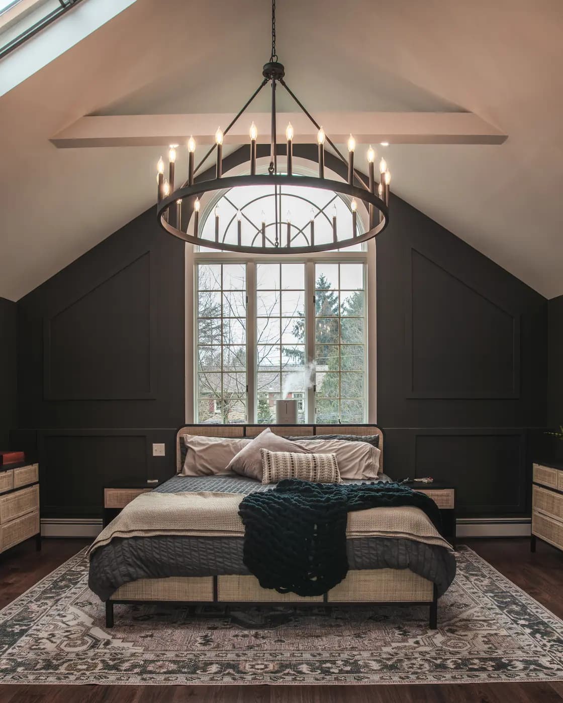 Dark moody bedroom with a vaulted ceiling, large arched window, and round chandelier
